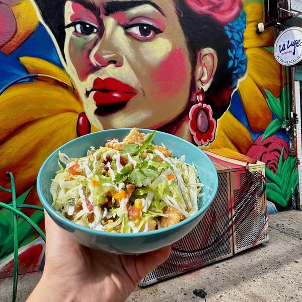 A hand holds a bowl of La Lupe Cantina's Mexican salad in front of a colorful mural.
